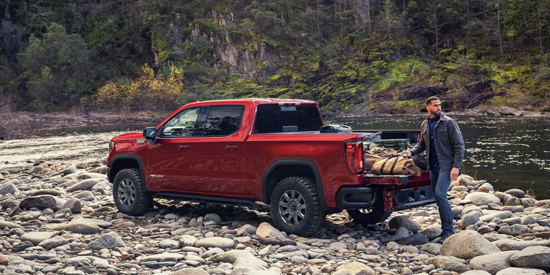 A red 2026 GMC Sierra 1500 is parked out in the open in the mountains on a very rocky shore beside a lazy mountain stream. The owner is lifting camping equipment out of the trunk of the truck.