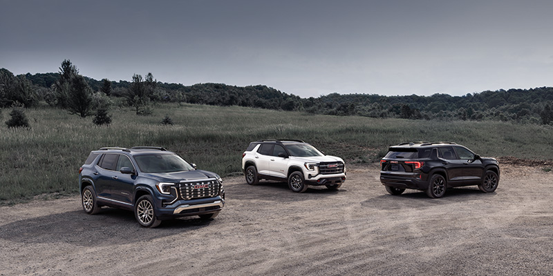 A grey, a white and a black 2026 GMC Terrain are parked on a sandy lot in the middle of a country field with low wooded hills in the ba
