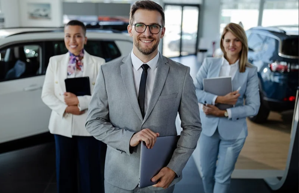 2 women and a male salespersons are holding their Ipads and smiling at us on the sunlit floor of a GMC sales floor with vehicles on display behind them.